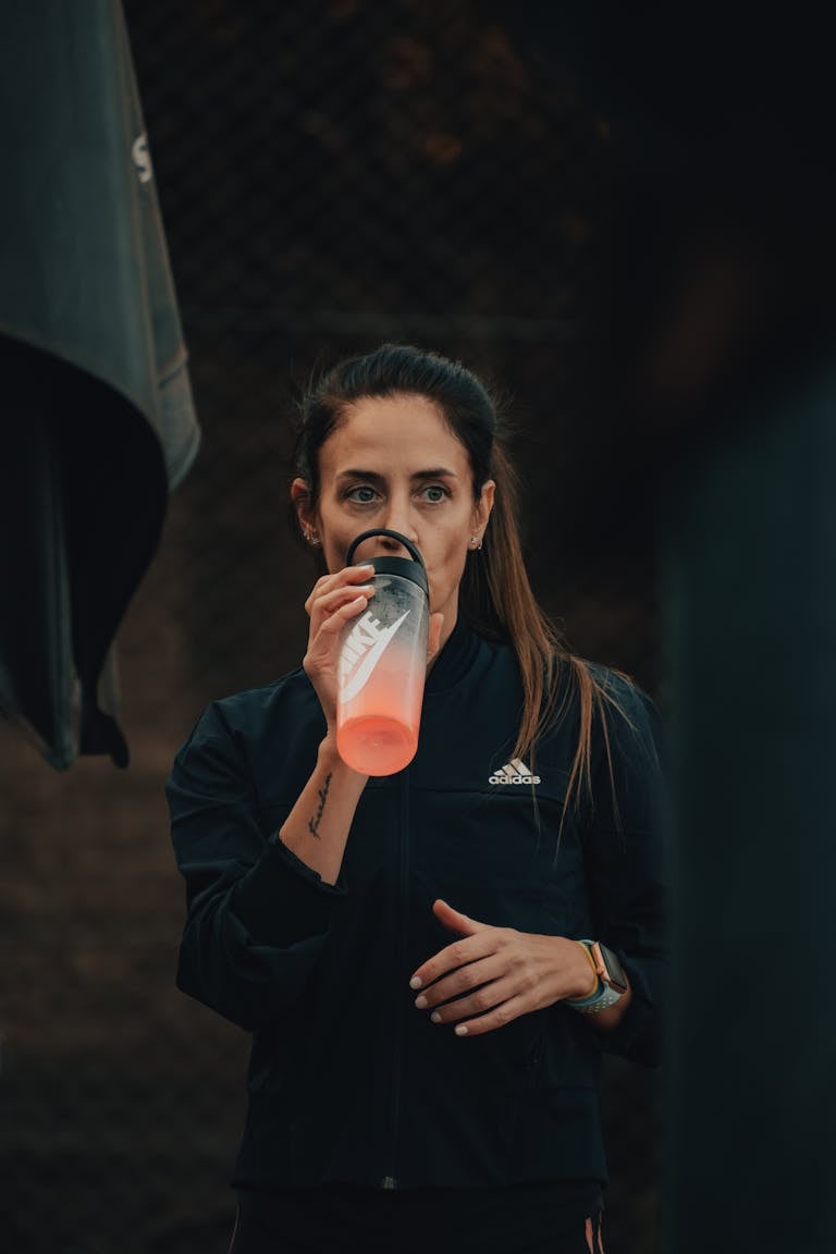 A woman in a black jacket hydrates with a tumbler while exercising outdoors.