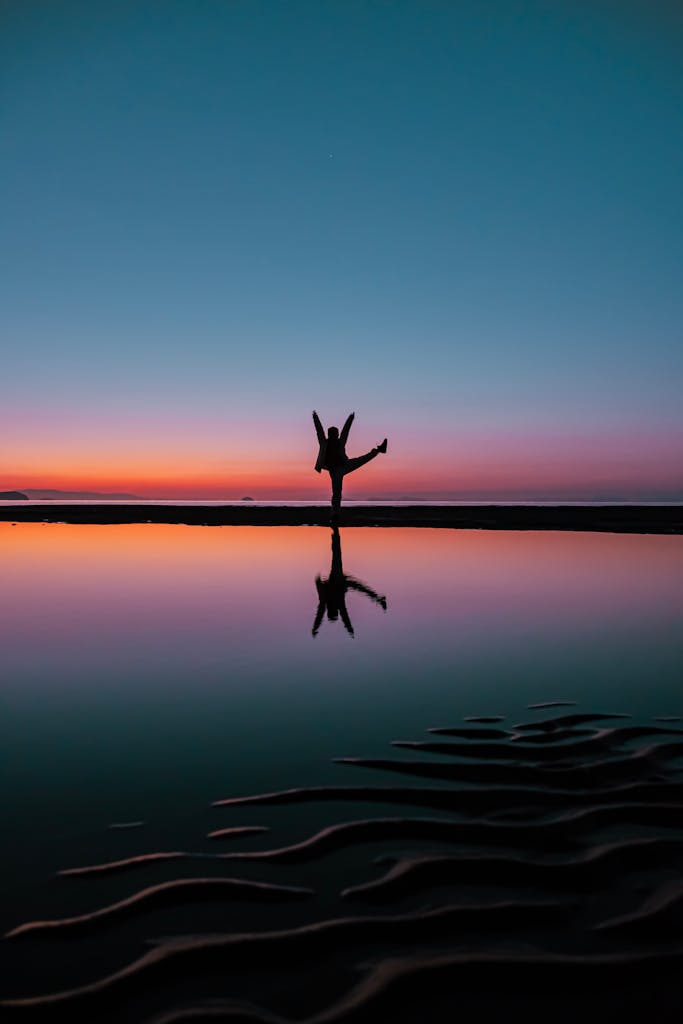 A person poses with arms raised at twilight, reflected on calm water at the sea shore.