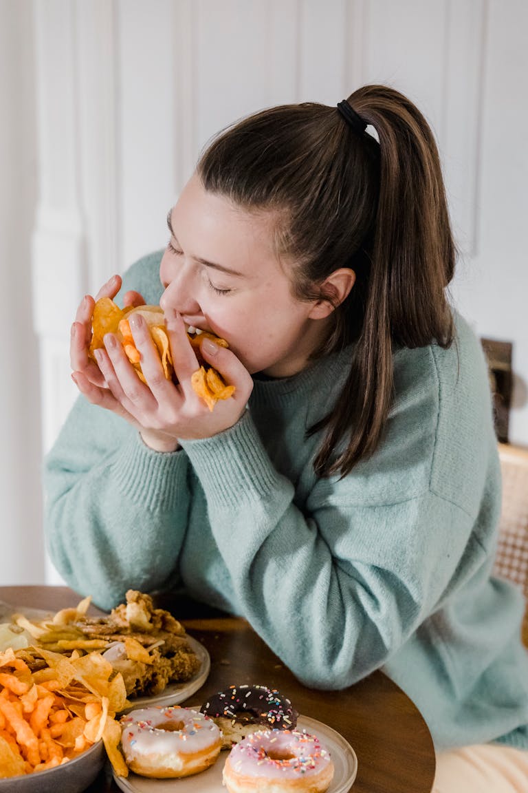 Hungry female in casual clothes eating crispy chips from hands while sitting at table with assorted junk food at home